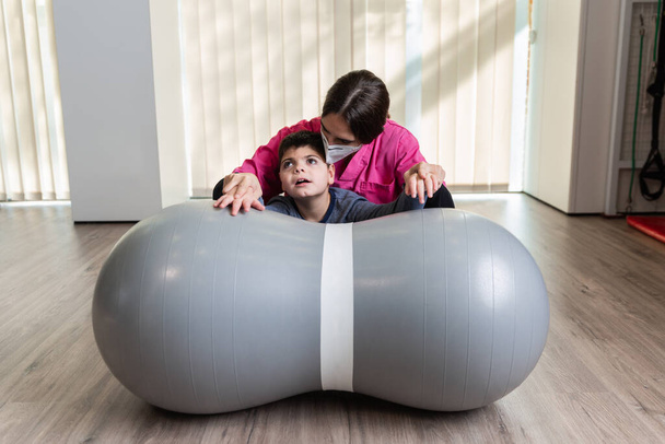 behindertes Kind und Physiotherapeut auf einem Peanut Gym Ball, der Gleichgewichtsübungen macht. Pandemieschutz - Foto, Bild