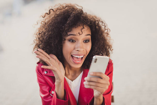 Photo of surprised funny curly woman dressed red jacked looking holding modern device rising hand arm outdoors urban city street - Фото, зображення