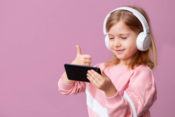 Little girl blogger influencer records videos from the blog on a smartphone, communicates with subscribers, puts likes, listens to music with headphones, remote learning on an isolated background. - Φωτογραφία, εικόνα