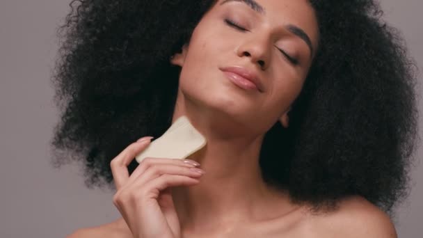 donna afroamericana utilizzando pietra di sollevamento su viso e collo isolato su grigio - Filmati, video