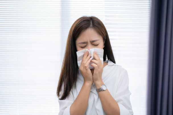 Asian woman sneezes at home. She uses a tissue to cover her mouth. - Foto, Imagem