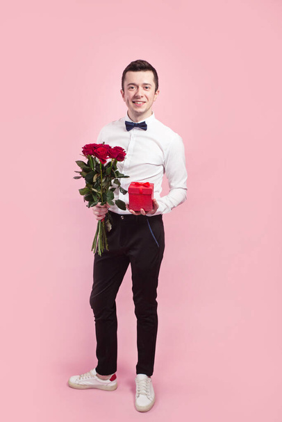Valentines day. Handsome smiing boy holding flowers and gift box on pink background. - Foto, Imagem