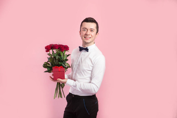 Ημέρα του Αγίου Βαλεντίνου. Όμορφος σιδηρουργός αγόρι κρατώντας λουλούδια και κουτί δώρου σε ροζ φόντο. - Φωτογραφία, εικόνα