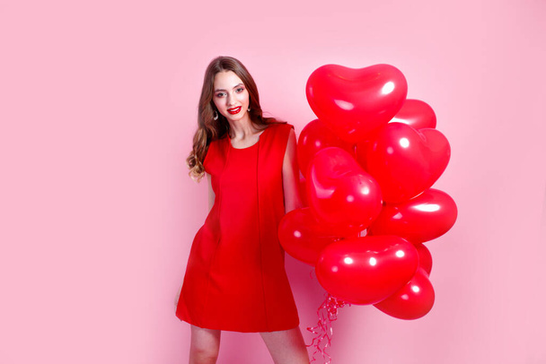 Valentines Day. Beautiful elegant young woman in red dress with heart shape red air balloons on pink background. - Foto, Imagem