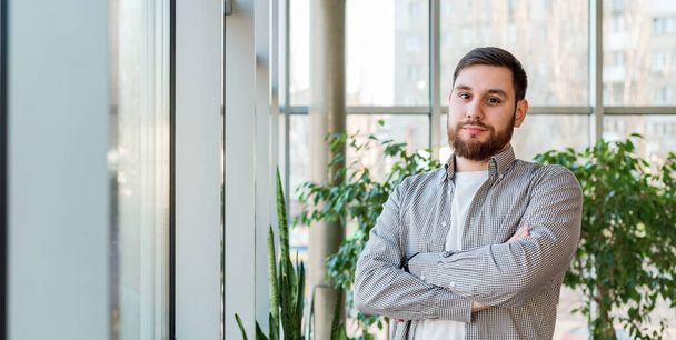 Caucasian man standing near full length window in modern office with plants. Successful bearded young man crossed arms. Handsome businessman smart man in casual shirt. Long web banner with copy space - Φωτογραφία, εικόνα