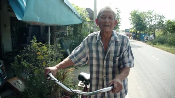 Scena al rallentatore di povero asiatico anziano dai capelli bianchi, più di settanta anni sorride e guida felicemente la bicicletta sul ciglio della strada del suo villaggio. - Filmati, video