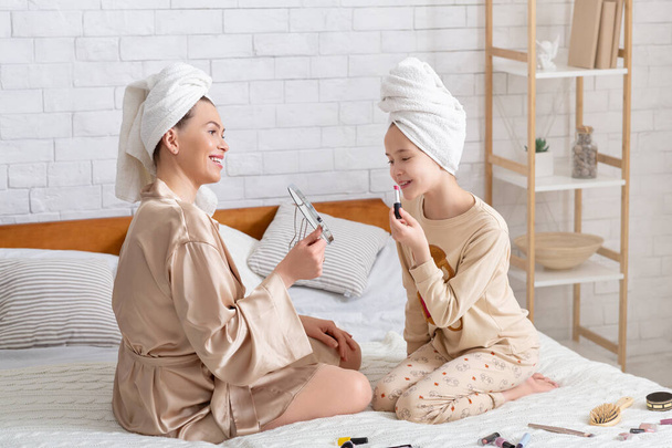 Belle femme et sa fille en vêtements de maison et serviettes de bain s'amuser avec le maquillage, l'application de rouge à lèvres sur le lit à la maison - Photo, image