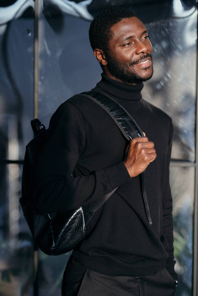 African american man in black trousers and shirt holding a bag on his back smiling - Foto, afbeelding