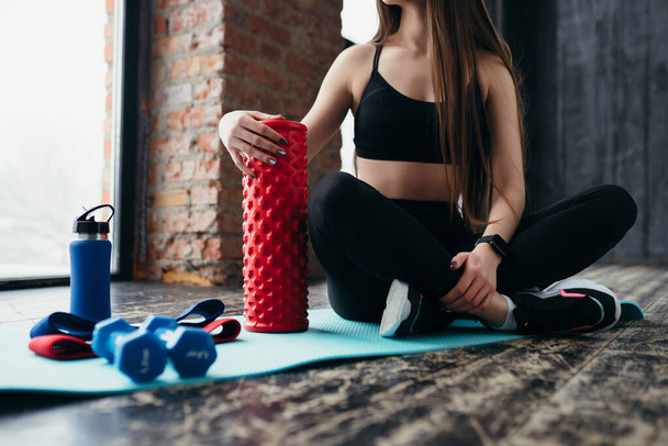 Trainer in a black top and leggings sits on a mat in the gym with a thermos and sports elastic bands - Fotografie, Obrázek