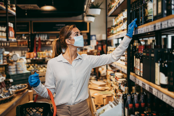 Beautiful young and elegant woman with face protective mask and gloves buying healthy food and drink in a modern supermarket or grocery store. Pandemic or epidemic lifestyle and consumerism concept. - Foto, afbeelding