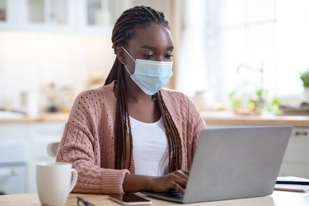 Молодая черная деловая женщина работает из дома с ноутбуком, носит медицинскую маску - Фото, изображение