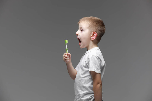 Ευτυχισμένο παιδί αγόρι με ηλεκτρική οδοντόβουρτσα σε γκρι φόντο. Υγειονομική περίθαλψη, οδοντιατρική υγιεινή. Mockup, αντίγραφο χώρου. - Φωτογραφία, εικόνα