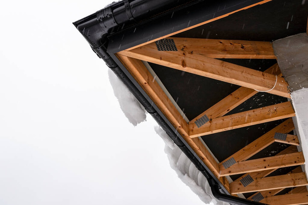 Крыша односемейного дома покрыта снегом, видны фермы крыши снизу и висячая шапка снега на канаве. - Фото, изображение