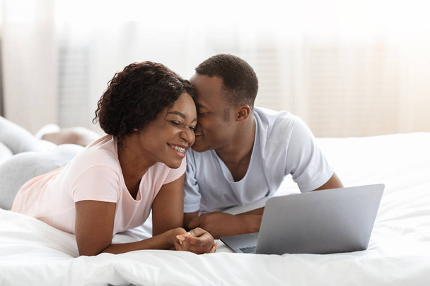 Νεαρό χαριτωμένο ζευγάρι της Αφρικής χρησιμοποιώντας φορητό υπολογιστή, ενώ στηρίζεται στο κρεβάτι - Φωτογραφία, εικόνα