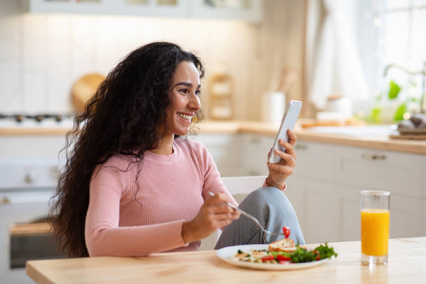 Onnellinen nuori nainen käyttää älypuhelinta keittiössä samalla kun maukas aamiainen - Valokuva, kuva