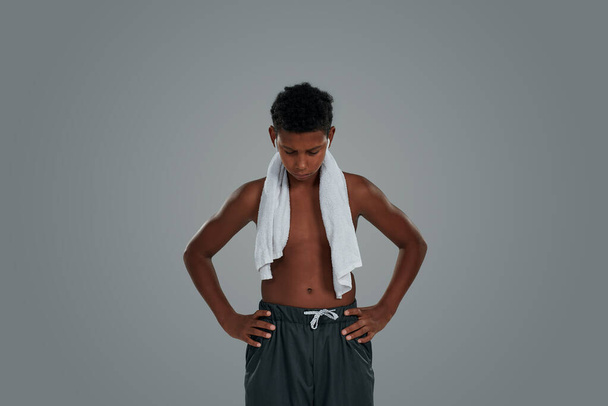 Feeling exausted after workout. Half naked teenage african boy with towel on shoulders keeping head down while standing against grey background - Фото, зображення
