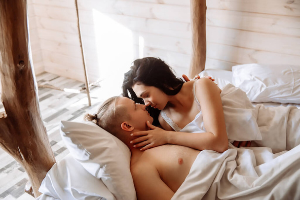 Νεαρό ζευγάρι ερωτευμένο, άντρας και γυναίκα, ξαπλωμένο στο κρεβάτι κάτω από μια λευκή κουβέρτα. Κοιτάζονται με τρυφερότητα.. - Φωτογραφία, εικόνα