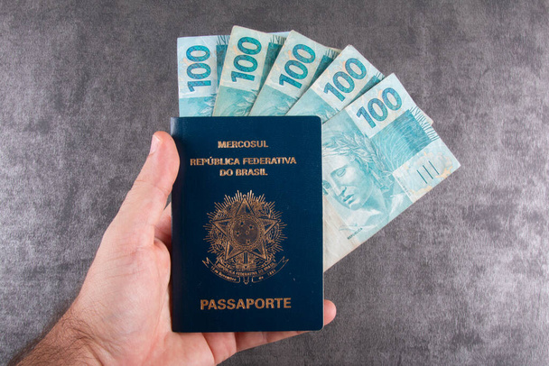 Hand hält brasilianischen Pass mit Hundert-Reais-Scheinen. - Foto, Bild