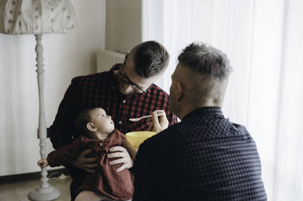 Coppia gay maschile con bambina adottata a casa - Due bei papà nutrono la bambina in cucina - Babysitter maschi - Famiglia Lgbt a casa - Concetto di diversità - Foto, immagini