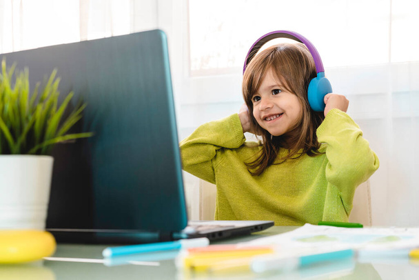 Grundschülerin mit Kopfhörer sitzt am Schreibtisch am Laptop - Lächelndes nettes Mädchen, das von zu Hause aus online lernt und während des Coronavirus-Ausbruchs im Internet lernt - Homeschooling-Konzept - Foto, Bild