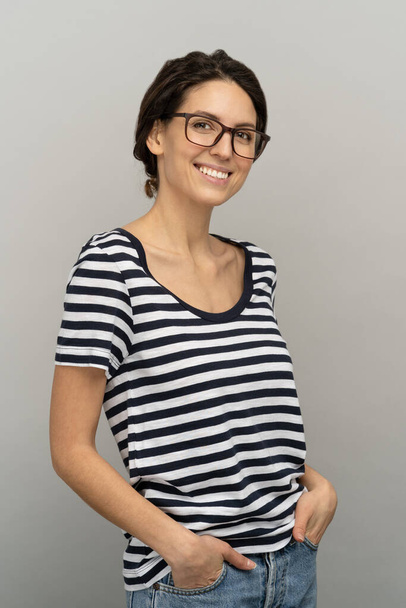 Улыбающаяся молодая европейская женщина в раздетых футболках носит очки, стоя с руками в карманах - Фото, изображение