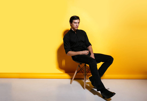 Retrato de comprimento total de um homem de moda sentado na cadeira olhando para a câmera, posando em estúdio, em fundo amarelo. Vista horizontal. - Foto, Imagem