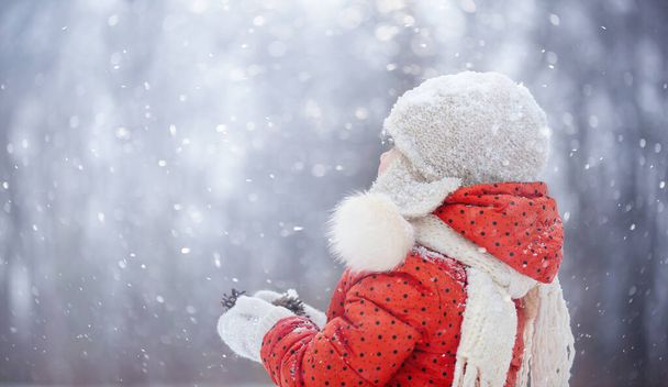 Ένα κοριτσάκι με πορτοκαλί μπουφάν κι ένα μάλλινο καπέλο περπατάει στο χιόνι μ 'ένα έλκηθρο κι ένα γλειφιτζούρι - Φωτογραφία, εικόνα