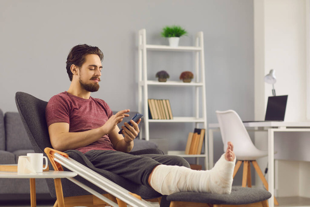 Young man with broken leg sitting in comfortable armchair and using mobile phone - Фото, изображение