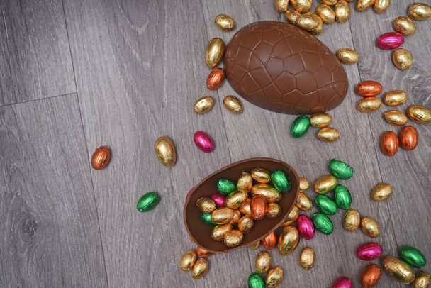 Hromada nebo skupina pestrobarevných a různých velikostí barevné fólie zabalené čokoládové velikonoční vejce v růžové, červené, zlaté a zelené. Velké poloviny hnědého mléka čokoládové vejce mají mini vejce uvnitř, na šedém dřevěném pozadí.  - Fotografie, Obrázek