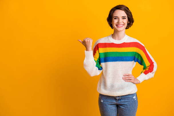 Photo of funky cute brunette hair girl stand pointing empty space wear rainbow sweater denim jeans isolated on bright yellow background - Photo, Image