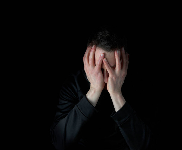 Hombre maduro deprimido con la cara hacia abajo en ambas manos mientras está rodeado de oscuridad  - Foto, imagen
