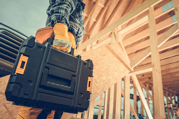 Construction Contractor with Plastic Power Tools Box in His Hand Going to Wooden Skeleton Frame Construction Site of House. Industrial Theme. - Photo, image