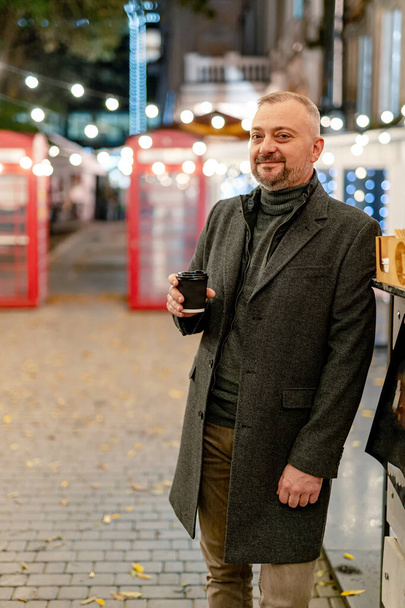 Half body portrait of a happy businessman standing outdoors with cup of coffee. Elegant coat and suit. Modern building background. - Фото, изображение