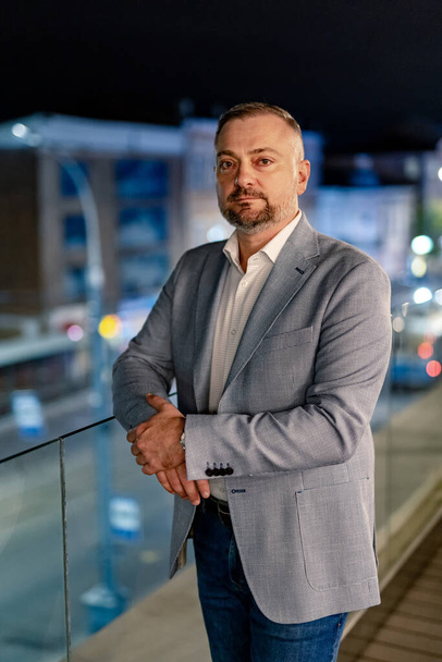Mature businessman on a hotel balcony. Evening panoramic view on a city. Man in grey jacket. Blurred background. - Fotografie, Obrázek