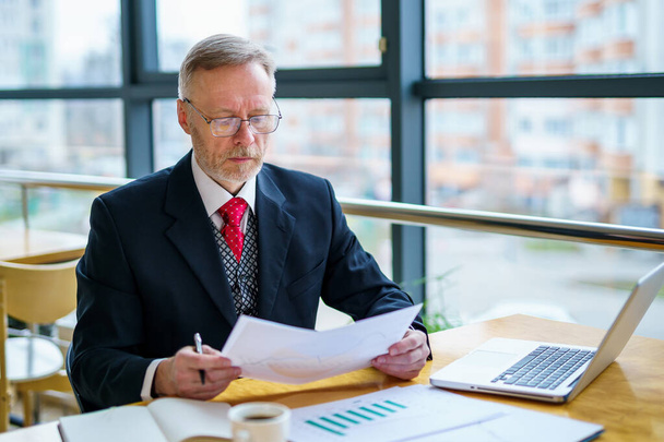 Thoughtful middle aged businessman in suit with a laptop on table while working with documents. Red tie and dark suit. - Foto, Imagem