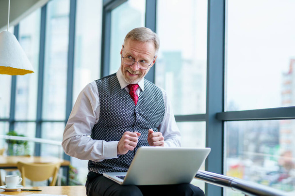 Cheerful mature businessman smiles to the camera with laptop on knees. Male in white shirt, grey vest and red tie. Big windows on the background. - Valokuva, kuva