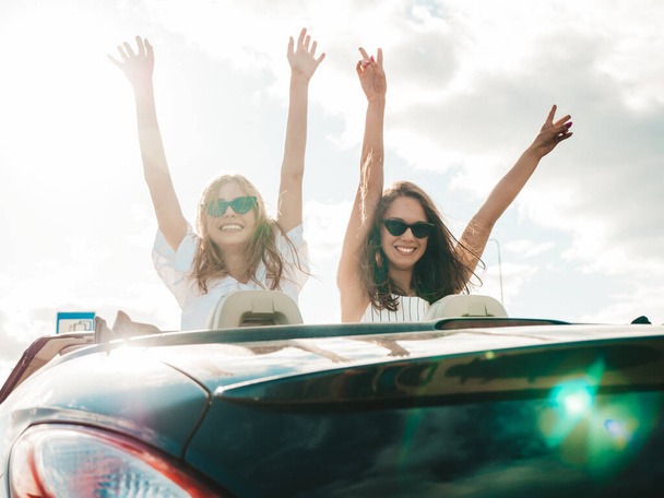 Portrait of two young beautiful and smiling hipster female in convertible car. Sexy carefree women driving cabriolet. Positive models riding and having fun in sunglasses. They raising hands - Φωτογραφία, εικόνα