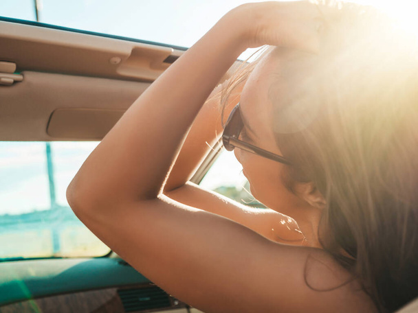 Portrait of young beautiful and smiling hipster female in convertible car. Sexy carefree woman driving cabriolet. Positive model riding and having fun in sunglasses at sunset - Foto, imagen