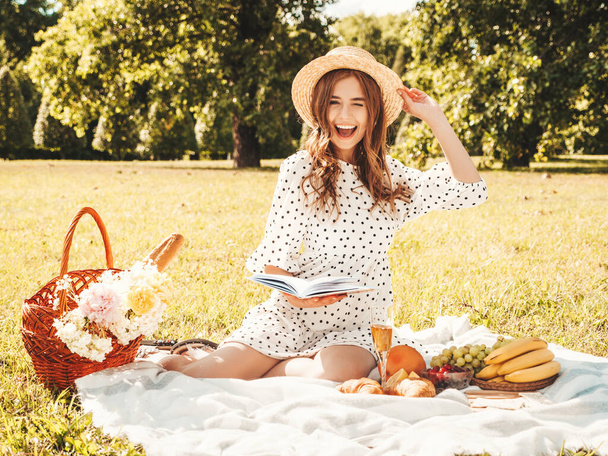 Young beautiful hipster woman in trendy summer sundress and hat. Carefree woman making picnic outside. Positive model sitting on plaid on grass, eating fruits and cheese, reading book - Fotó, kép