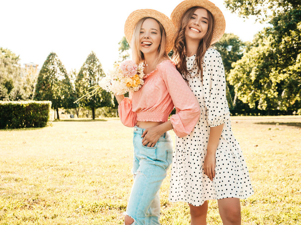 Two young beautiful smiling hipster woman in trendy summer sundress. Sexy carefree women posing in the park in hats. Positive models having fun and hugging at sunset. They holding flowers - Foto, Imagem