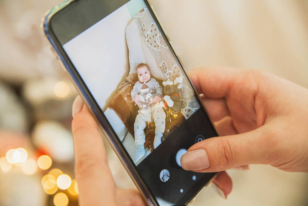 Мама фотографирует по телефону ребенка с игрушкой и гирляндой на диване - Фото, изображение