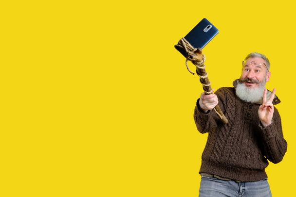 Studio portret van volwassen man met baard selfie met houten stok, grappige selfie maken concept, gele achtergrond - Foto, afbeelding