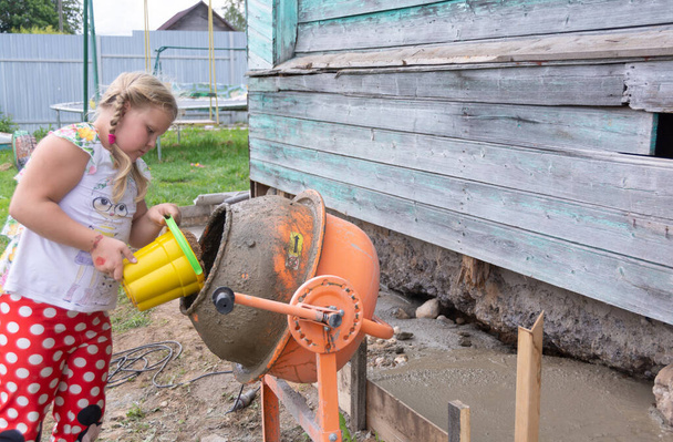 A little helper. In the village, a girl helps her father, pours water into a concrete mixer. - Photo, image