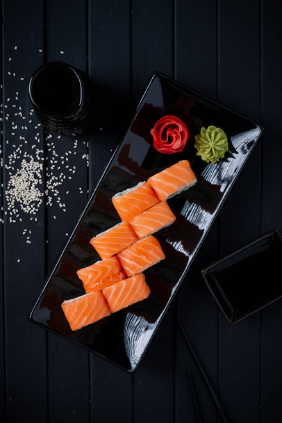 Philadelphia rolka set s lososem, okurkou a smetanovým sýrem na japonském talíři na černém dřevěném pozadí s hůlkami a sójovou omáčkou - Fotografie, Obrázek