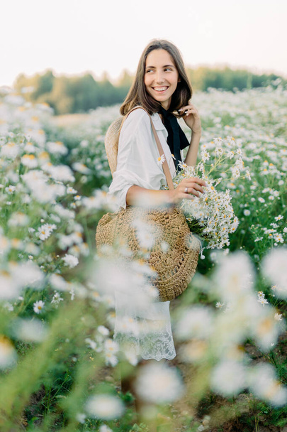 attractive beautiful young brunette girl in a white dress, with straw bag and hat in the middle of a field of chamomile daisies. Blurred background and selective focus. - Photo, image