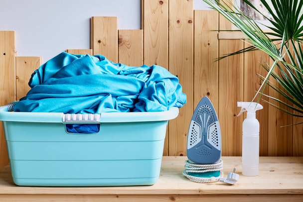 Basket with clean laundry, iron and spray bottle on table at home. Real laundry room with wooden wall. Household concept - Photo, Image