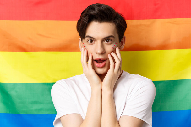 Primer plano de la hermosa morena gay hombre en camiseta blanca, con maquillaje brillo, mirando divertido a la cámara, tocando la cara, de pie contra la bandera del orgullo del arco iris - Foto, imagen