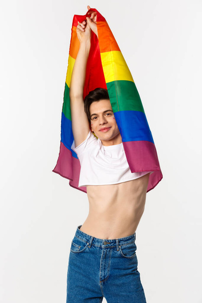 Vista verticale di bello androgino gay uomo alzando bandiera arcobaleno e sorridente felice, in piedi in crop top e jeans su sfondo bianco - Foto, immagini