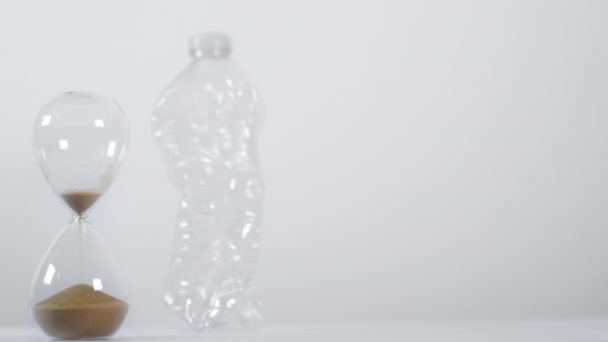 A wide shot of an emptying sand hourglass next to a crushed single-use plastic bottle on a white background - Footage, Video