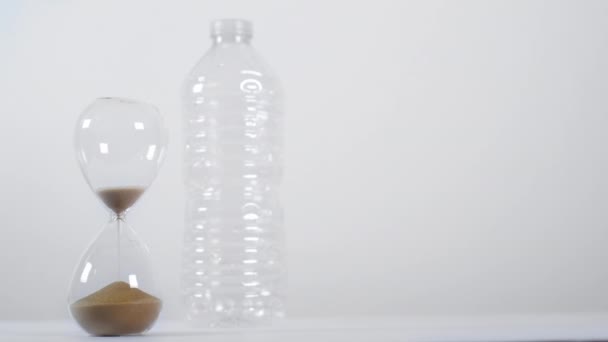 Vue d'ensemble d'un sablier vide à côté d'une bouteille en plastique à usage unique sur fond blanc - Séquence, vidéo